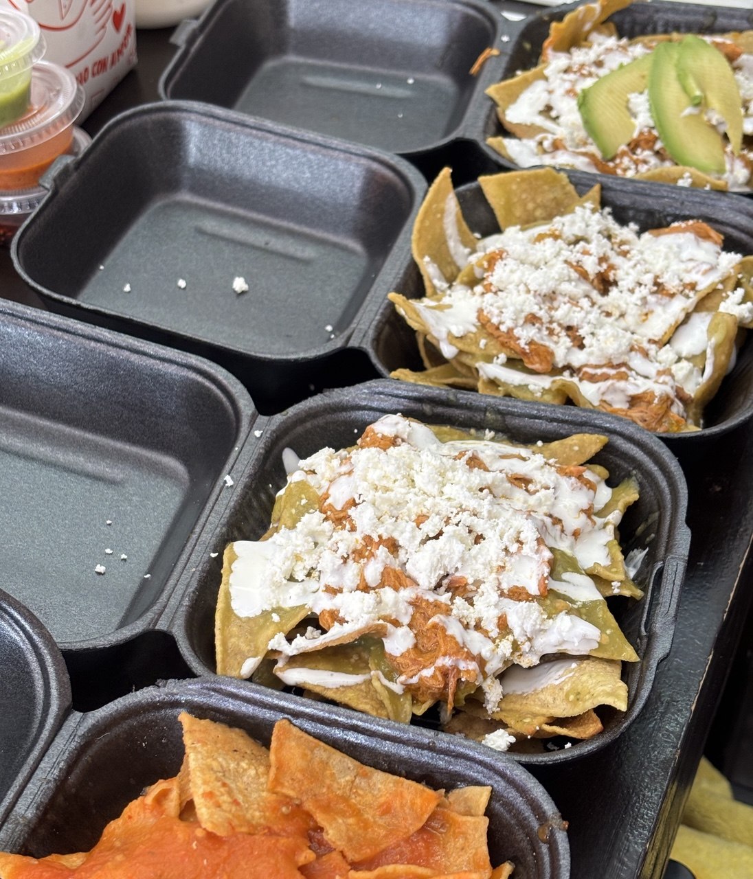 Chilaquiles in black takeout boxes: green and red salsa, crema, queso fresco, avocado.